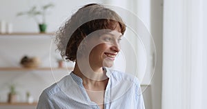 Middle aged optimistic dreamy woman standing near window looking outside