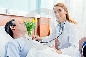 Middle aged patient lying on bed and doctor examining him with stethoscope