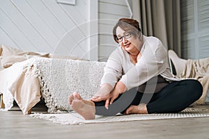 Middle aged plus size smiling brunette woman in home wear doing stretching in bedroom at home