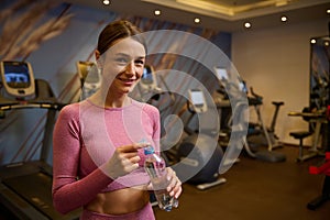 Middle aged pretty woman with a bottle of water, looking at camera standing at gym after workout. Healthy lifestyle, fitness,