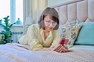 Middle aged relaxed woman lying on bed at home using smartphone