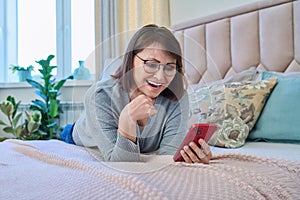 Middle aged relaxed woman lying on bed at home using smartphone