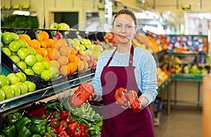 Middle-aged saleswoman holding peppers at food stall in greengrocery