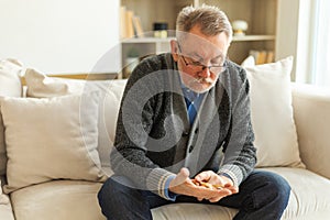 Middle aged senior man holding medical pills sitting on couch at home. Mature old senior grandfather taking medication