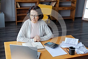 Middle aged senior woman sit with laptop and paper document, Pensive older mature lady reading paper bill pay online at