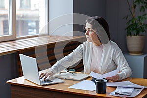 Middle aged senior woman sit with laptop and paper document, Pensive older mature lady reading paper bill pay online at