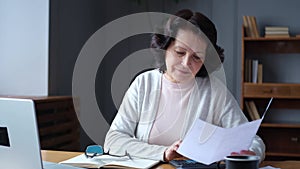 Middle aged senior woman sit with laptop and paper document, Pensive older mature lady reading paper bill pay online at