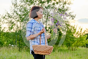 Middle-aged smiling woman with a basket and a bouquet of wildflowers in nature