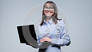 Middle aged smiling woman holding laptop in hands on gray background