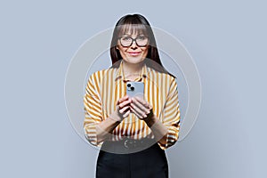 Middle aged smiling woman with smartphone on grey studio background