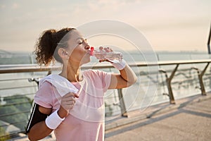 Middle-aged sporty woman in a pink T-shirt and gray leggings, with white wristbands, holds a terry towel on her shoulders and