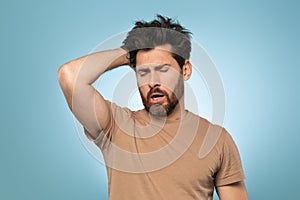 Middle aged tired and very sleepy man keeping hand on head and yawning, standing over blue studio background, free space