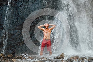 Middle-aged topless sincerely laughing man standing under the mountain river waterfall and enjoying the splashing Nature power.