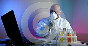 A middle-aged virologist is in the lab, sitting at a table with a laptop, flasks of tests on the table, examining