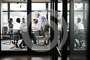 Middle aged white businessman stands addressing colleagues at meeting, seen through glass wall