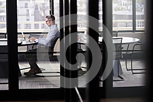 Middle aged white businessman working alone in a meeting room, seen through glass door