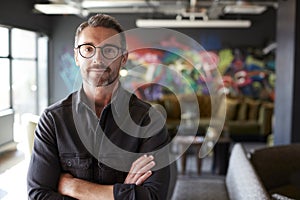 Middle aged white male creative in casual office lounge area looks to camera, arms crossed, waist up