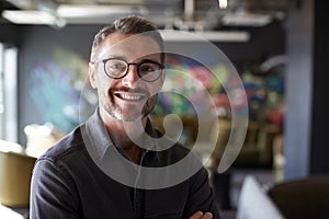 Middle aged white male creative in casual office lounge area looks to camera smiling, close up