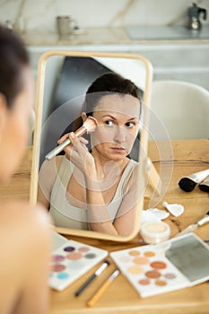 Middle-aged woman applying blush on her cheeks by means of makeup brush