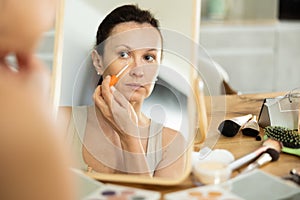 Middle-aged woman applying concealer on her facial skin sitting in front of the mirror