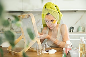 Middle-aged woman applying eye patch mask on her face sitting in front of the mirror