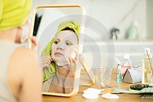 Middle-aged woman applying tissue mask on her facial skin sitting in front of the mirror