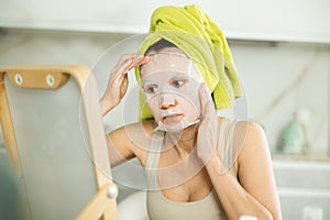 Middle-aged woman applying tissue mask on her facial skin sitting in front of the mirror