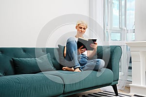 A middle aged woman with blond hair is relaxing on a sofa in her living room, reading a novel