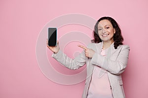Middle-aged woman in casual gray jacket, smiling and showing a smartphone with blank screen, isolated on pink background