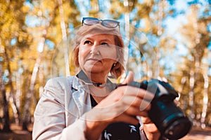 Middle-aged woman checking images on camera in autumn forest. Senior woman walking and enjoying hobby