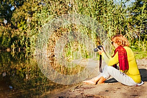 Middle-aged woman checking images on camera sitting by autumn river bank. Senior woman enjoying hobby