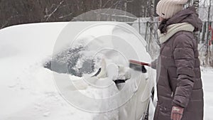 A middle-aged woman cleans car windows from snow in the winter season. Snow drifts
