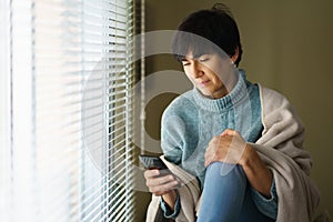 Middle-aged woman consulting something on her smartphone near the window at home.