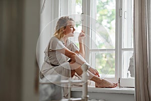 A middle-aged woman in a cream dress sits mysteriously and looks out the window on the windowsill. Green trees outside.