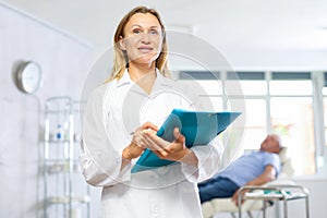 Middle-aged woman doctor posing in doctor's cabinet with document in hands