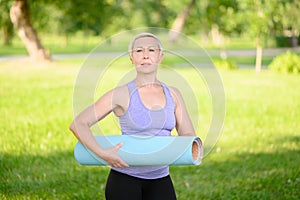 Middle-aged woman doing smiling and holding a fitness mat outside, leading a healthy lifestyle.Time to sport.