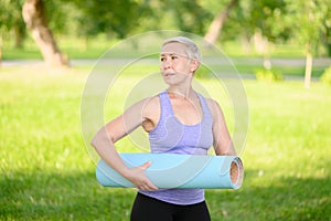 Middle-aged woman doing smiling and holding a fitness mat outside, leading a healthy lifestyle.Time to sport.