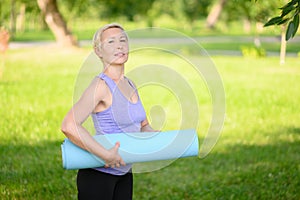 Middle-aged woman doing smiling and holding a fitness mat outside, leading a healthy lifestyle.Time to sport.