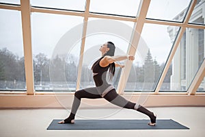 Middle aged woman doing yoga in Virabhadrasana One or Warrior One yoga pose on the mat in front of large windows., exercise fitnes