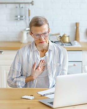 Middle aged woman explaining symptoms to doctor online while sitting in kitchen at home
