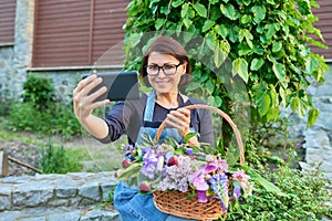 Middle aged woman in garden with basket of fresh spring flowers taking selfie photo