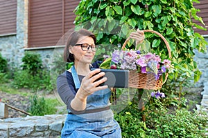 Middle aged woman in garden with basket of fresh spring flowers taking selfie photo