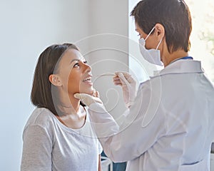 Middle-aged woman general practitioner in face protective mask examining patient throat in clinic