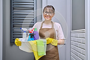 Middle-aged woman in gloves apron with basin of detergents in bathroom