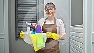 Middle-aged woman in gloves apron with basin of detergents in bathroom
