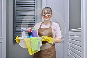 Middle-aged woman in gloves apron with basin of detergents in bathroom