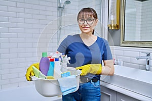 Middle-aged woman in gloves with basin of detergents in bathroom