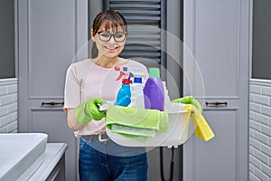 Middle-aged woman in gloves with basin of detergents in bathroom