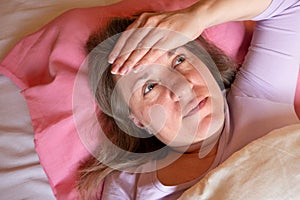 Middle aged woman has a high temperature. 40 years women lying in the bed and touches the forehead with hand. She is unwell and