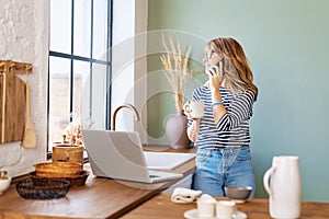 Middle aged woman having a phone call and using laptop while standing at the kitchen at home